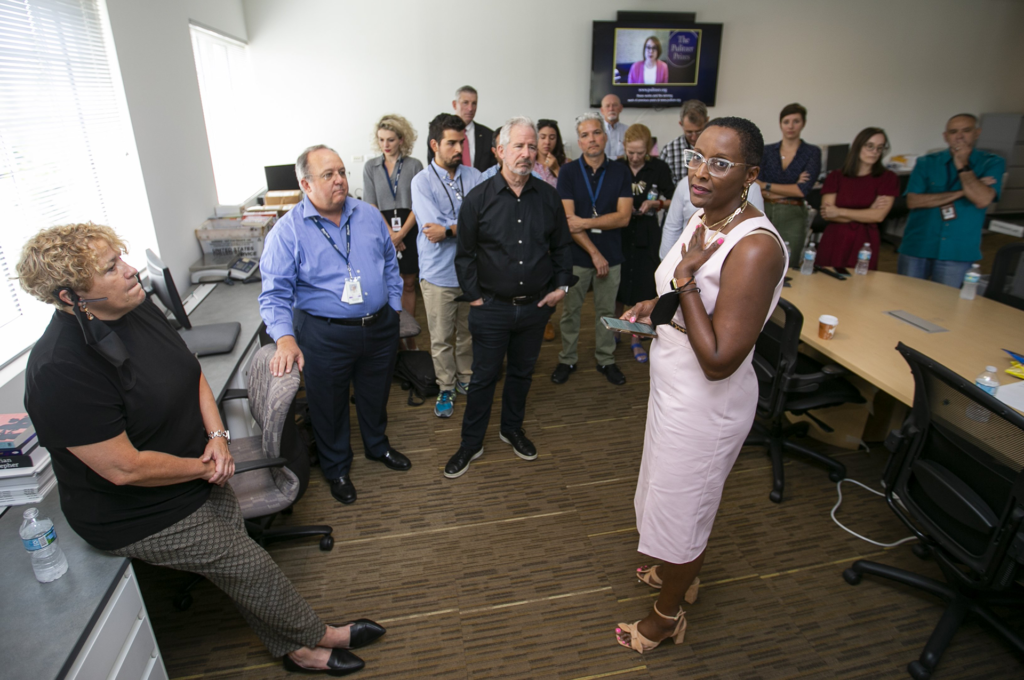 A woman in glasses and a pink dress stands in the center of a group of people in an officer conference room who are looking at her, as she looks at the camera. In the background, a television screen shows the words, “The Pulitzer Prize.”