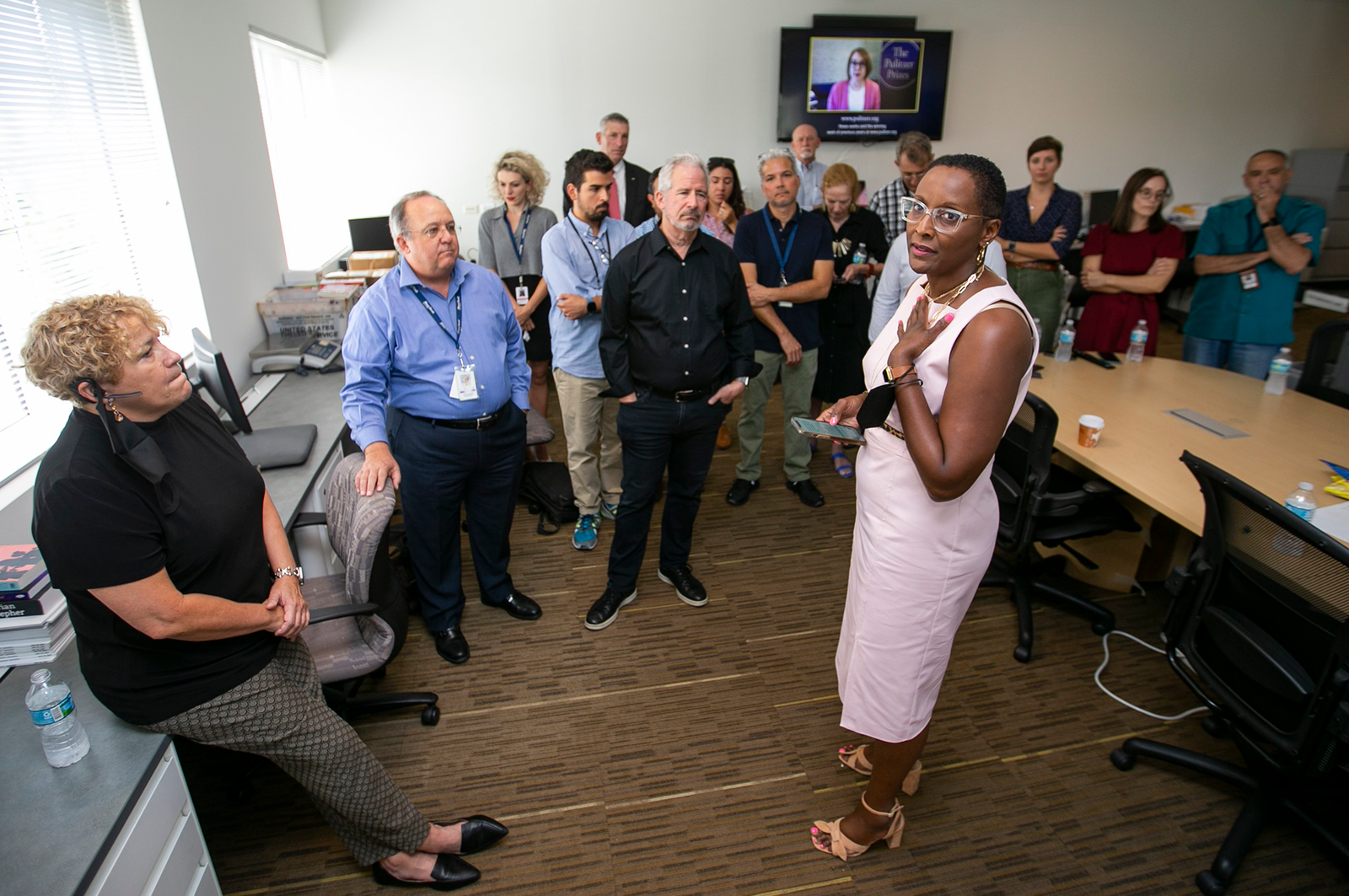 A woman in glasses and a pink dress stands in the center of a group of people in an officer conference room who are looking at her, as she looks at the camera. In the background, a television screen shows the words, “The Pulitzer Prize.”